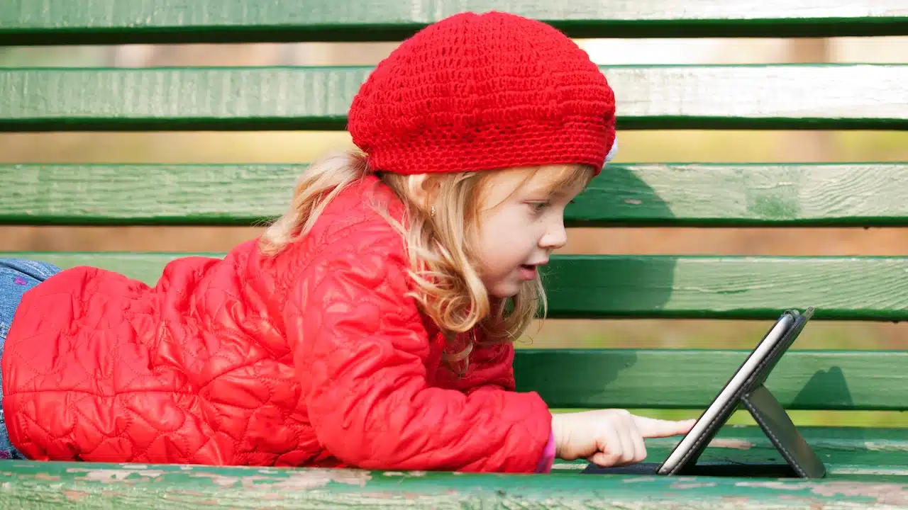 Happy smiling little girl working with tablet pc and lying on bench in the park.