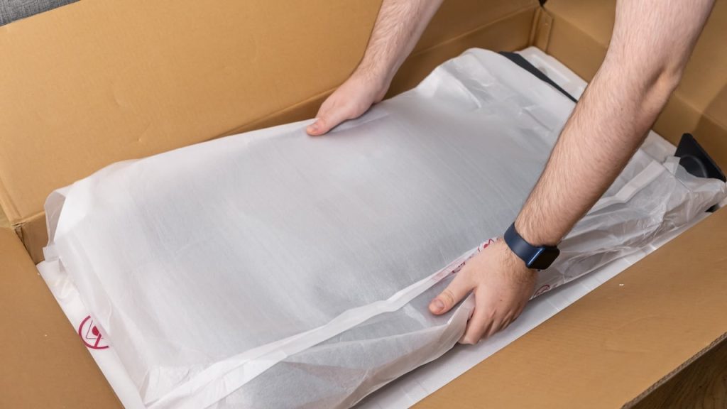 View of human hands carefully taking a new electronic item in its white protective packaging from a brown cardboard delivery box, showcasing online commerce