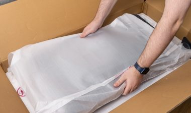 View of human hands carefully taking a new electronic item in its white protective packaging from a brown cardboard delivery box, showcasing online commerce