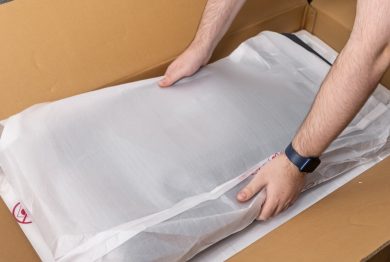 View of human hands carefully taking a new electronic item in its white protective packaging from a brown cardboard delivery box, showcasing online commerce