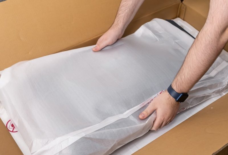 View of human hands carefully taking a new electronic item in its white protective packaging from a brown cardboard delivery box, showcasing online commerce