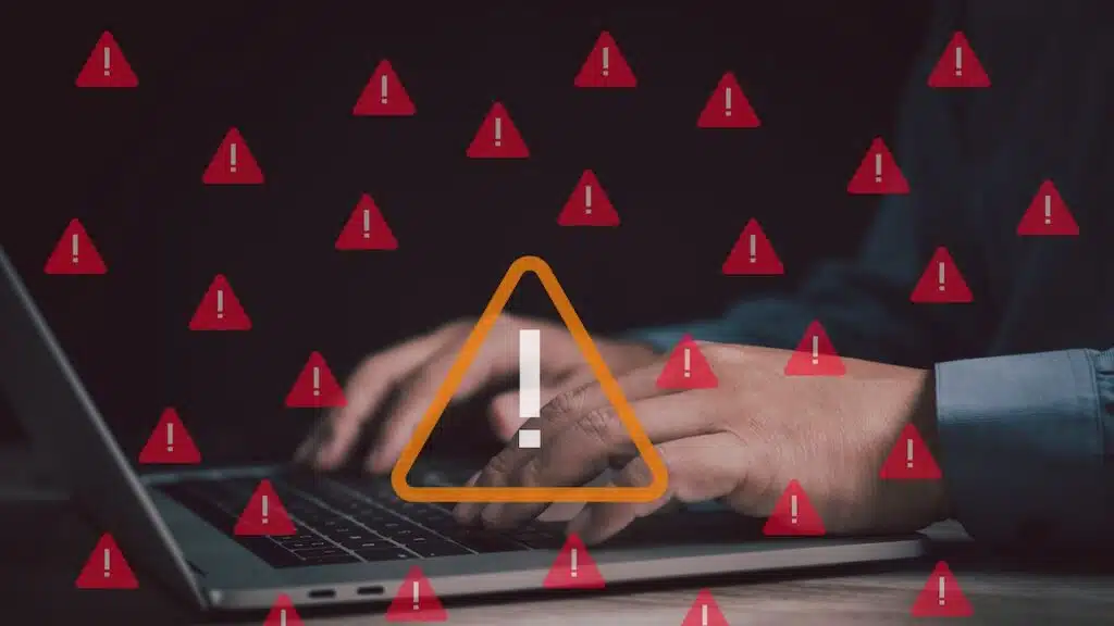 man works on a laptop surrounded by red warning icons, symbolizing cybersecurity threats
