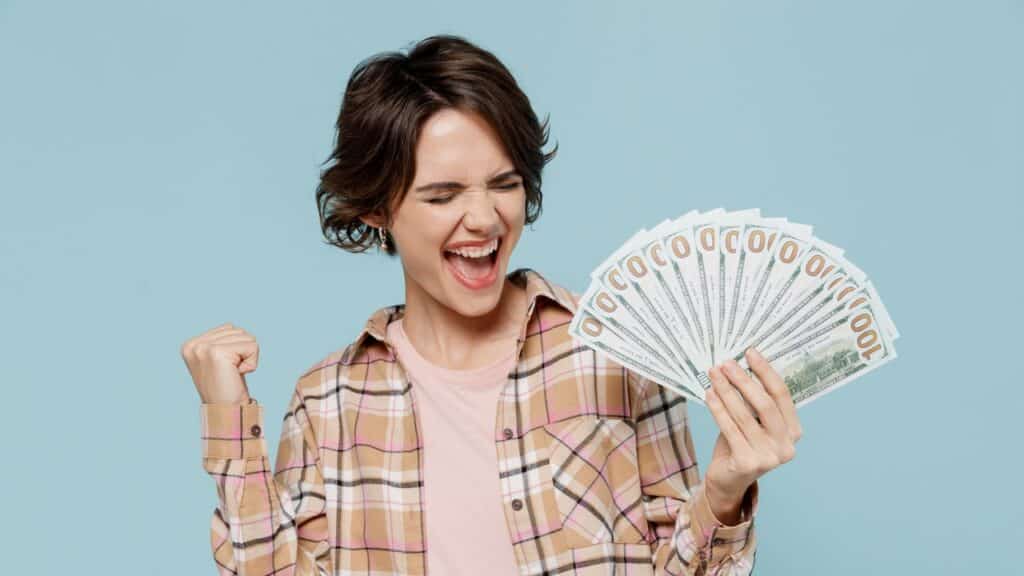 Young happy overjoyed excited woman 20s wearing casual brown shirt holding fan of cash money in dollar banknotes