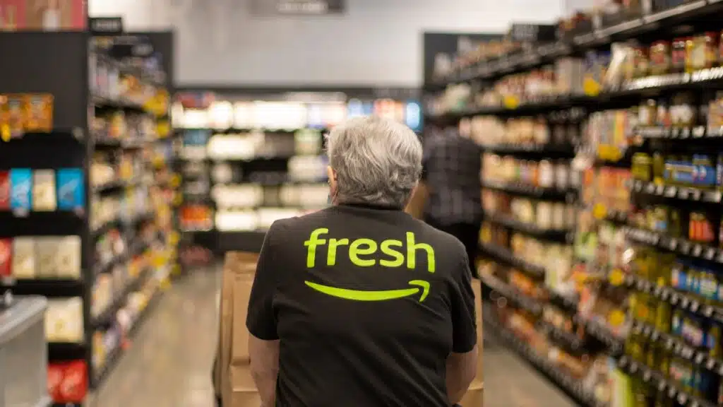 An elderly worker in an Amazon Fresh T-shirt pushes a cart loaded with pickup paper bags down the aisles inside an Amazon Fresh grocery store in Irvine, California.