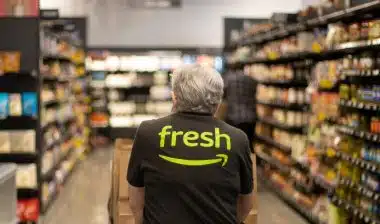 An elderly worker in an Amazon Fresh T-shirt pushes a cart loaded with pickup paper bags down the aisles inside an Amazon Fresh grocery store in Irvine, California.