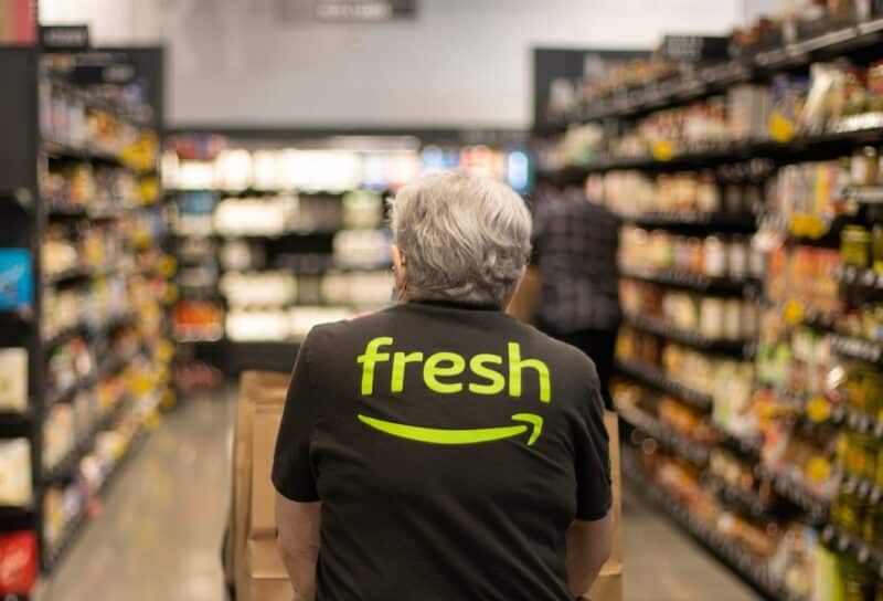 An elderly worker in an Amazon Fresh T-shirt pushes a cart loaded with pickup paper bags down the aisles inside an Amazon Fresh grocery store in Irvine, California.