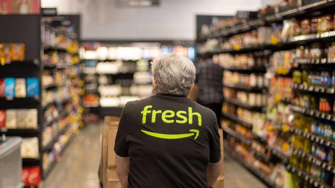 An elderly worker in an Amazon Fresh T-shirt pushes a cart loaded with pickup paper bags down the aisles inside an Amazon Fresh grocery store in Irvine, California.