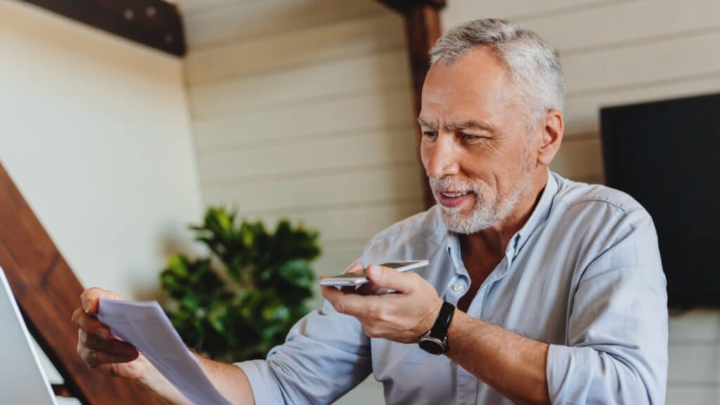Mature senior using mobile phone while working on laptop computer at home office.