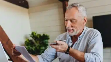 Mature senior using mobile phone while working on laptop computer at home office.