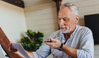 Mature senior using mobile phone while working on laptop computer at home office.