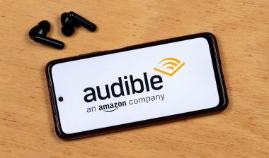 photo of a smartphone screen showing Audible app black logo with a wireless earphones over a wooden background