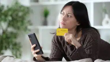 Worried asian woman thinking before online payment holding phone and credit card looking to the side