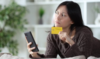 Worried asian woman thinking before online payment holding phone and credit card looking to the side