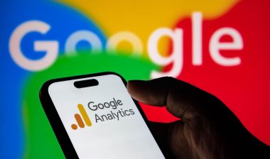 Closeup of a hand holding a smartphone displaying the google analytics logo with the google logo in the background
