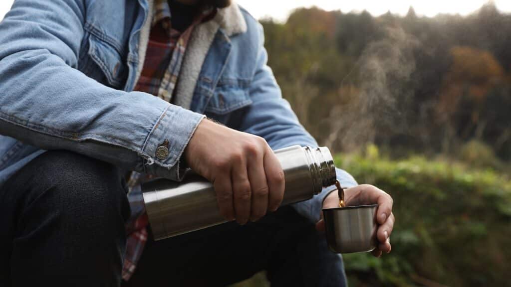 Man pouring hot drink from metallic thermos into cup lid in nature, closeup