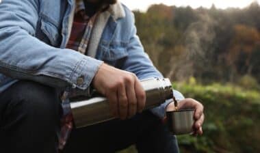 Man pouring hot drink from metallic thermos into cup lid in nature, closeup