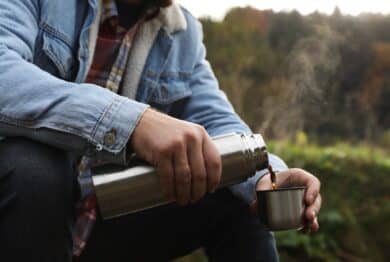 Man pouring hot drink from metallic thermos into cup lid in nature, closeup