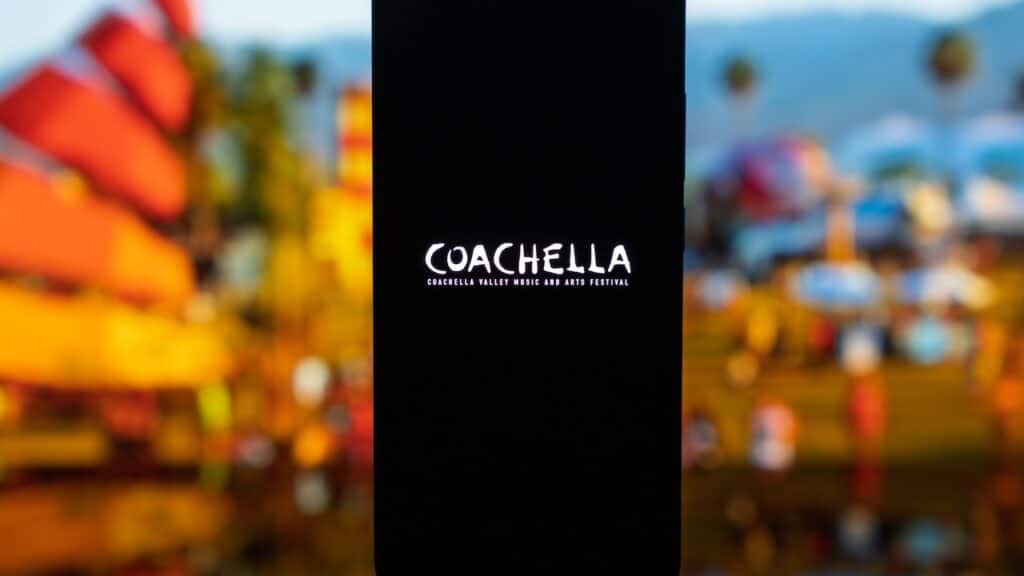 Smartphone displaying the Coachella Valley Music and Arts Festival logo on a dark screen, with a blurred outdoor festival background featuring palm trees and desert landscape, representing music culture and live events.