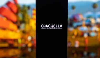 Smartphone displaying the Coachella Valley Music and Arts Festival logo on a dark screen, with a blurred outdoor festival background featuring palm trees and desert landscape, representing music culture and live events.