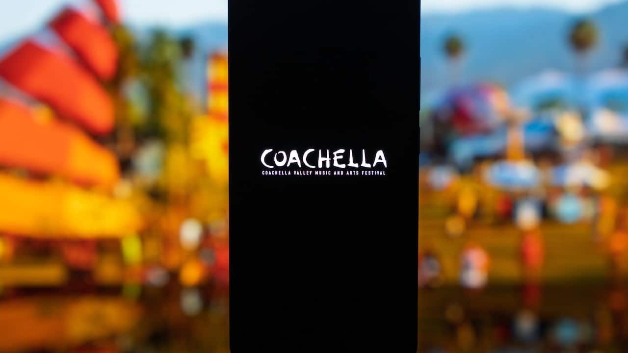 Smartphone displaying the Coachella Valley Music and Arts Festival logo on a dark screen, with a blurred outdoor festival background featuring palm trees and desert landscape, representing music culture and live events.