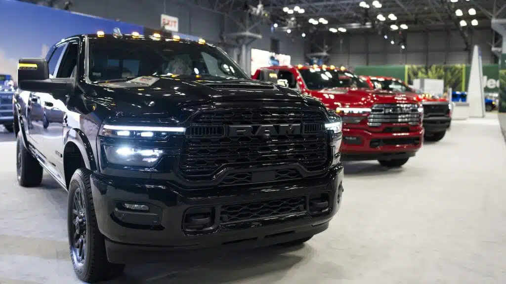 Black Ram pickup truck at 125th NY Auto Show Javits Center. Two red Ram trucks partially visible brightly lit large exhibition hall rugged design performance indoor display.