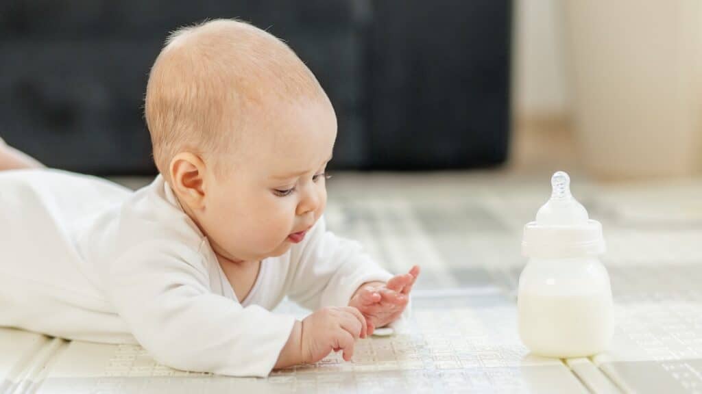 Curious baby in white clothes lying on tummy, exploring surroundings while baby formula bottle on floor