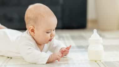 Curious baby in white clothes lying on tummy, exploring surroundings while baby formula bottle on floor
