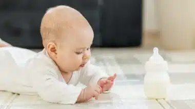 Curious baby in white clothes lying on tummy, exploring surroundings while baby formula bottle on floor