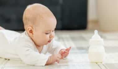 Curious baby in white clothes lying on tummy, exploring surroundings while baby formula bottle on floor