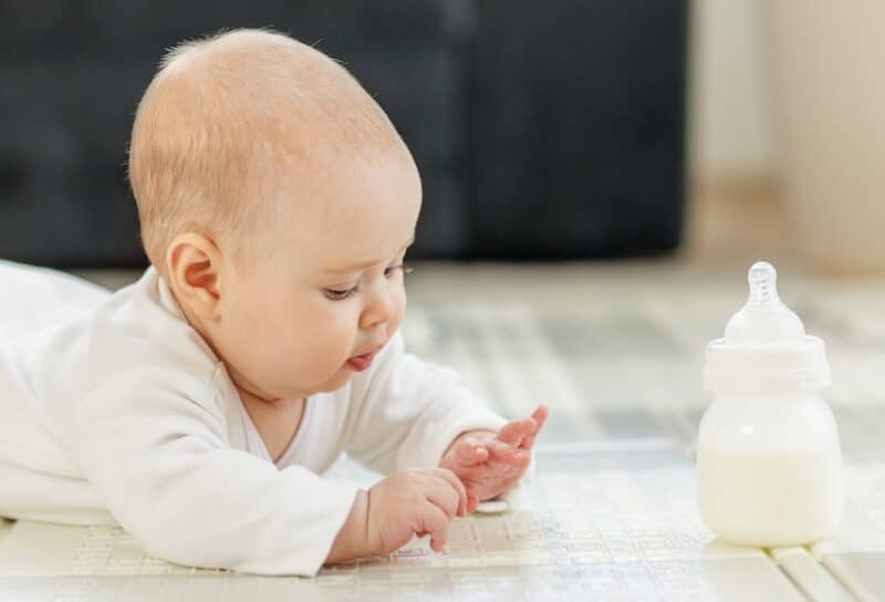 Curious baby in white clothes lying on tummy, exploring surroundings while baby formula bottle on floor