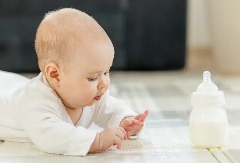 Curious baby in white clothes lying on tummy, exploring surroundings while baby formula bottle on floor