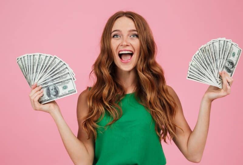 Portrait of a happy excited woman with long hair holding bunch of money banknotes in each hand