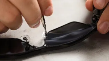 Handyman repairing sunglasses with screwdriver at grey table, closeup