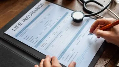 Close-up of hands filling out a life insurance form on a clipboard, with a stethoscope and medicine bottles on a wooden table.
