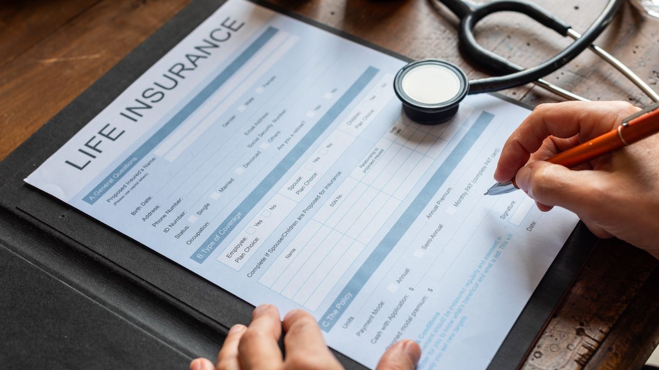 Close-up of hands filling out a life insurance form on a clipboard, with a stethoscope and medicine bottles on a wooden table.