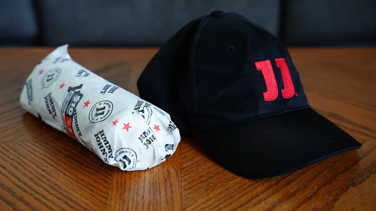 Sub sandwich wrapped in Jimmy John's food paper wrap sits on the table with employee hat.